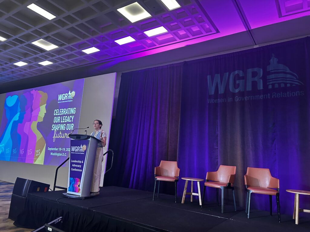 A woman speaks at a podium on a stage with purple lighting at the WGR conference, hosted by Beekeeper Group. Behind her, a screen displays "Celebrating Our Legacy, Shaping Our Future"—a fitting theme for this public affairs event. Three empty chairs are on stage.