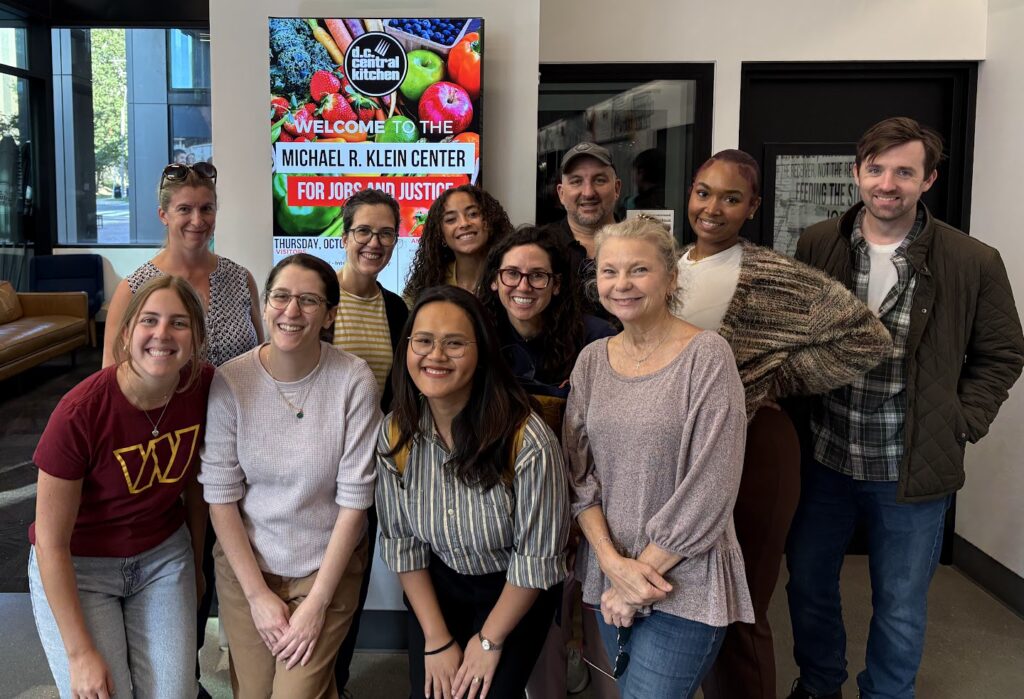 A group of twelve people from the Beekeeper Group smiles and poses together indoors in front of a sign that reads “Welcome to the Michael R. Klein Center for Jobs and Justice.” The public affairs atmosphere appears friendly and casual.