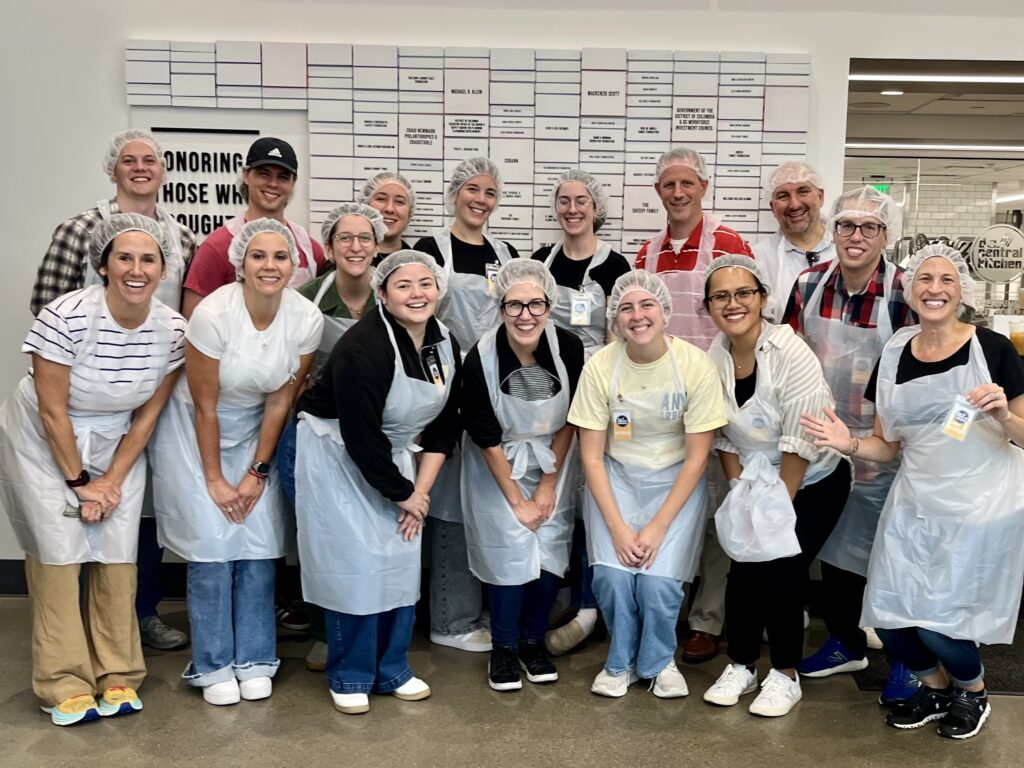A group of people wearing hairnets, aprons, and casual clothes pose and smile together indoors in front of a wall with a whiteboard displaying names and information, representing Beekeeper Group’s commitment to public affairs teamwork.