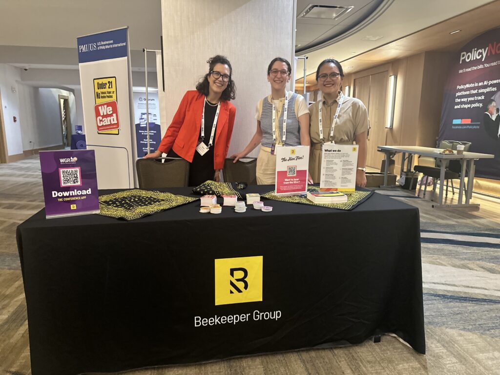 Three smiling people stand behind a table with a yellow Beekeeper Group sign, displaying informational materials and promo items at the Leadership & Advocacy Conference in a modern venue.