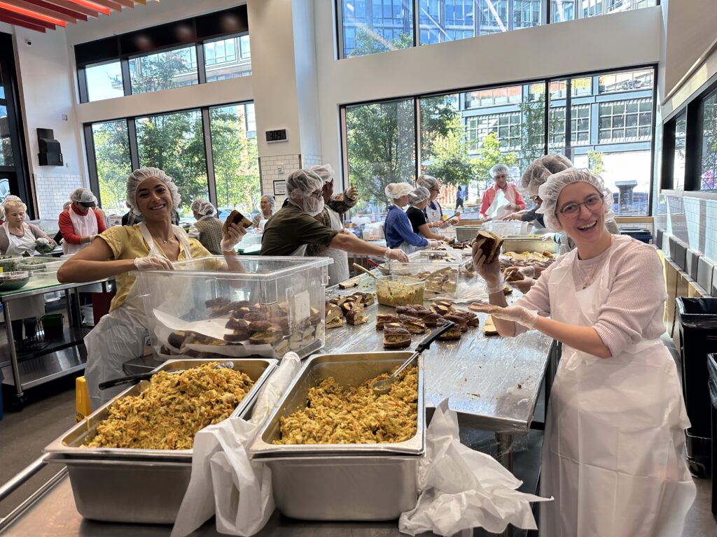 IMG_6354 Volunteers from the Beekeeper Group, wearing hairnets and aprons, prepare and pack food in a bright, modern kitchen with large windows. Trays of food and stacks of sandwiches fill the counter as everyone stays happy and busy.