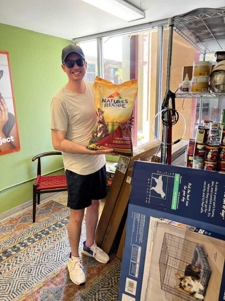 A smiling person in sunglasses and a cap holds a large bag of Nature’s Recipe dog food inside a pet store, surrounded by pet supplies, while representing Beekeeper Group’s public affairs team.