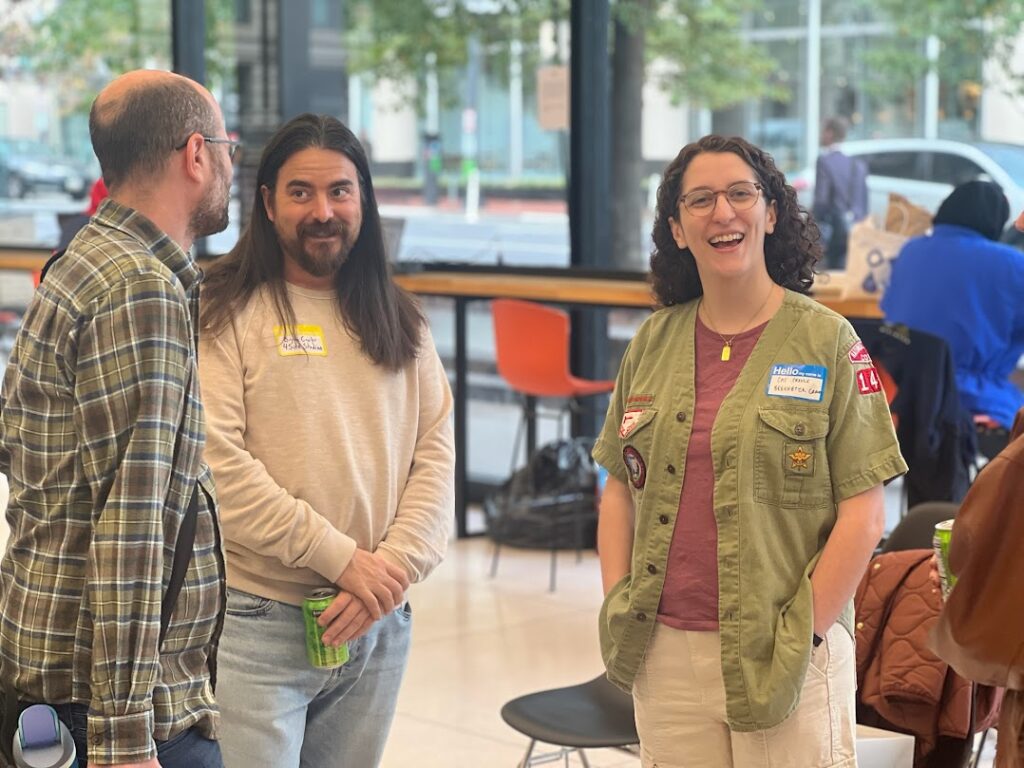 Three people stand indoors, relaxed and friendly, recapping Beekeeper Group's 2025 day of purpose. One wears glasses and a green jacket with patches, another has long hair and a beige sweater, and the third wears glasses and a plaid shirt.