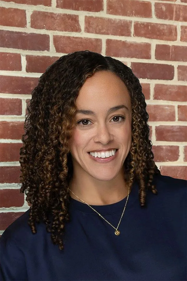 A woman with curly brown hair and a navy blue top smiles in front of a red brick wall, proudly celebrating 15 years with the Bees. She wears a simple gold necklace.