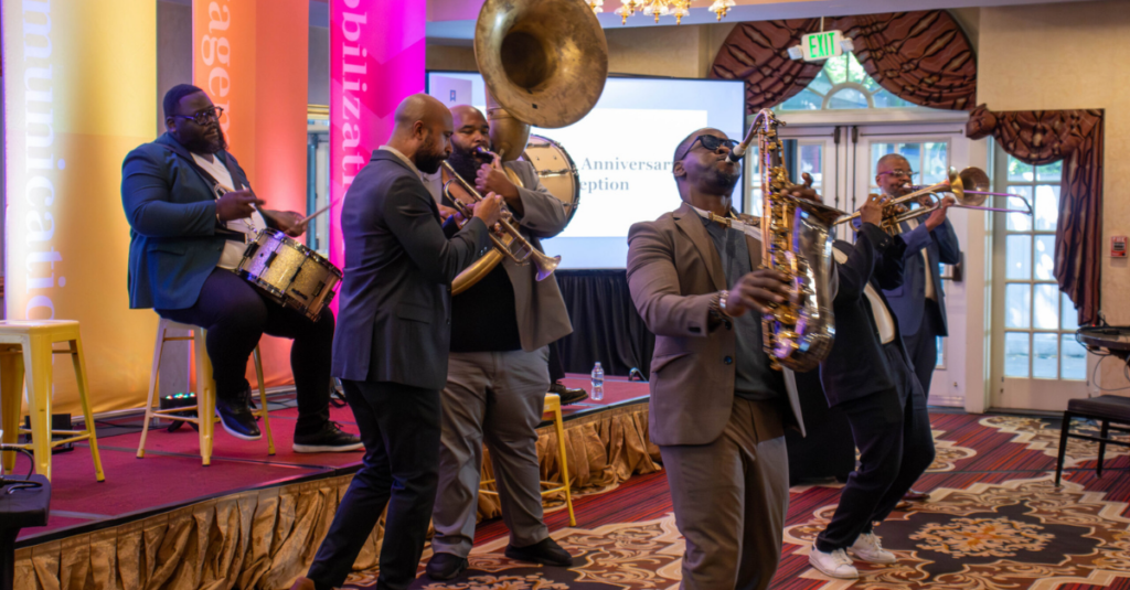 hive five graphics (8) A jazz band plays instruments on stage, including drums, trumpet, trombone, and saxophone, in front of a colorful backdrop and audience at an indoor event—perfect for featuring in the Summer 2026 edition of the Hive Five Newsletter.